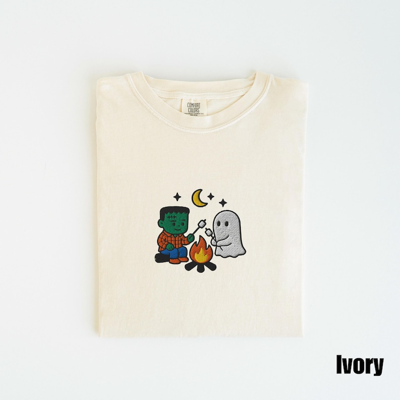 May include: Ivory-colored t-shirt with an embroidered design of a Frankenstein-like character and a ghost roasting marshmallows over a campfire under a crescent moon and stars. The shirt is folded neatly.