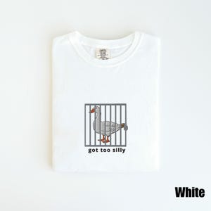 May include: White t-shirt featuring an embroidered goose inside a cage with the text "got too silly" below the image. The goose has gray feathers, an orange beak and feet. The shirt is labeled "Comfort Colors".