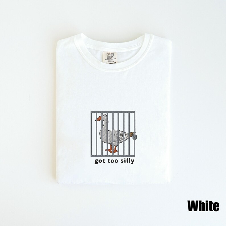 May include: White t-shirt featuring an embroidered goose inside a cage with the text "got too silly" below the image. The goose has gray feathers, an orange beak and feet. The shirt is labeled "Comfort Colors".
