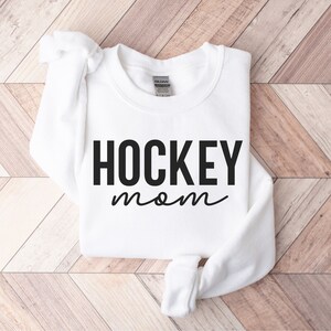 May include: White sweatshirt with black text that reads "HOCKEY mom" on a wooden background.
