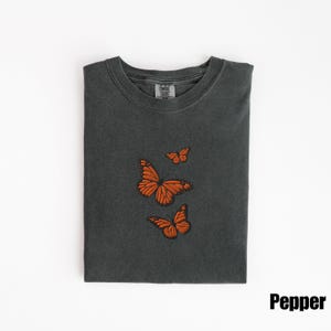 May include: A dark gray t-shirt with three embroidered orange butterflies. The butterflies are in different sizes and positions. The shirt is folded neatly on a white surface. The word "Pepper" is printed in black.