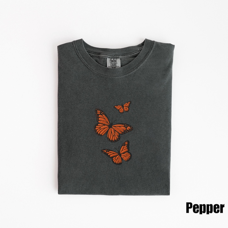 May include: A dark gray t-shirt with three embroidered orange butterflies. The butterflies are in different sizes and positions. The shirt is folded neatly on a white surface. The word "Pepper" is printed in black.