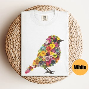 May include: White t-shirt with a floral bird design. The bird is made up of colorful flowers and is outlined in black. The shirt has a tag that says "Comfort Colors" on it.
