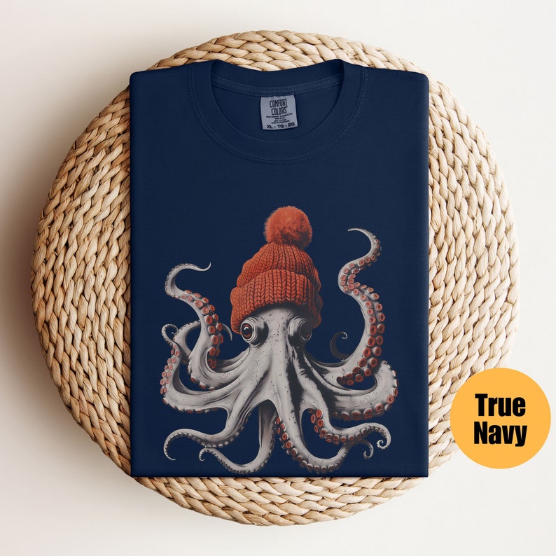 May include: A navy blue t-shirt featuring a detailed illustration of an octopus wearing an orange knit beanie with a pom-pom. The octopus has large eyes and red suckers on its tentacles. The shirt is folded on a woven surface.