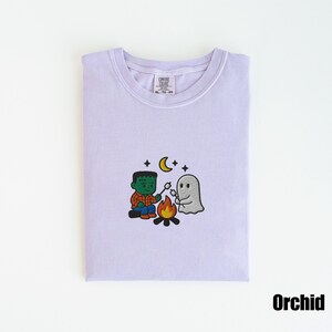 May include: Orchid-colored t-shirt with a crew neck. The shirt features an embroidered design of a Frankenstein-like character roasting marshmallows with a ghost over a campfire, under a crescent moon and stars.