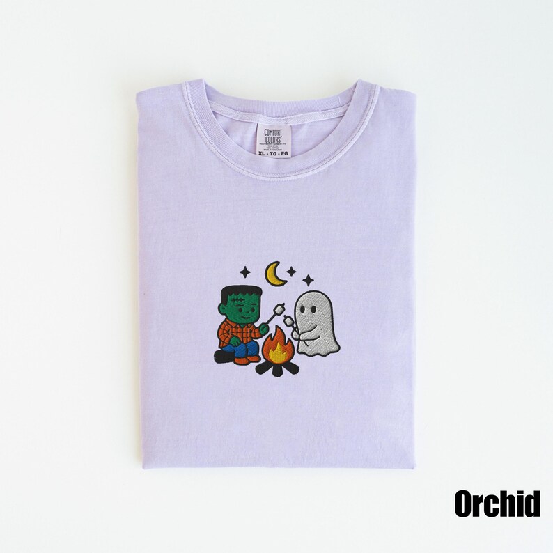May include: Orchid-colored t-shirt with a crew neck. The shirt features an embroidered design of a Frankenstein-like character roasting marshmallows with a ghost over a campfire, under a crescent moon and stars.
