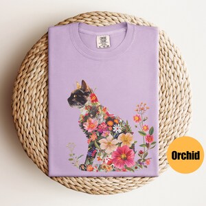 May include: A lavender t-shirt featuring a floral cat design. The cat is made up of pink, red, orange, and yellow flowers. The text "COMFORT COLORS" is visible on the tag.