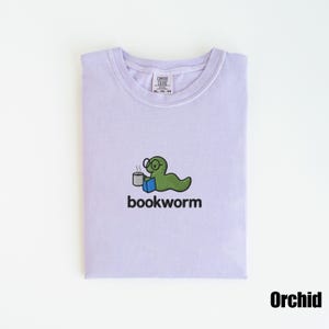 May include: A light purple t-shirt with a crew neck. The shirt features a green cartoon bookworm wearing glasses, holding a mug, and sitting on a blue book. The word "bookworm" is printed below the graphic.