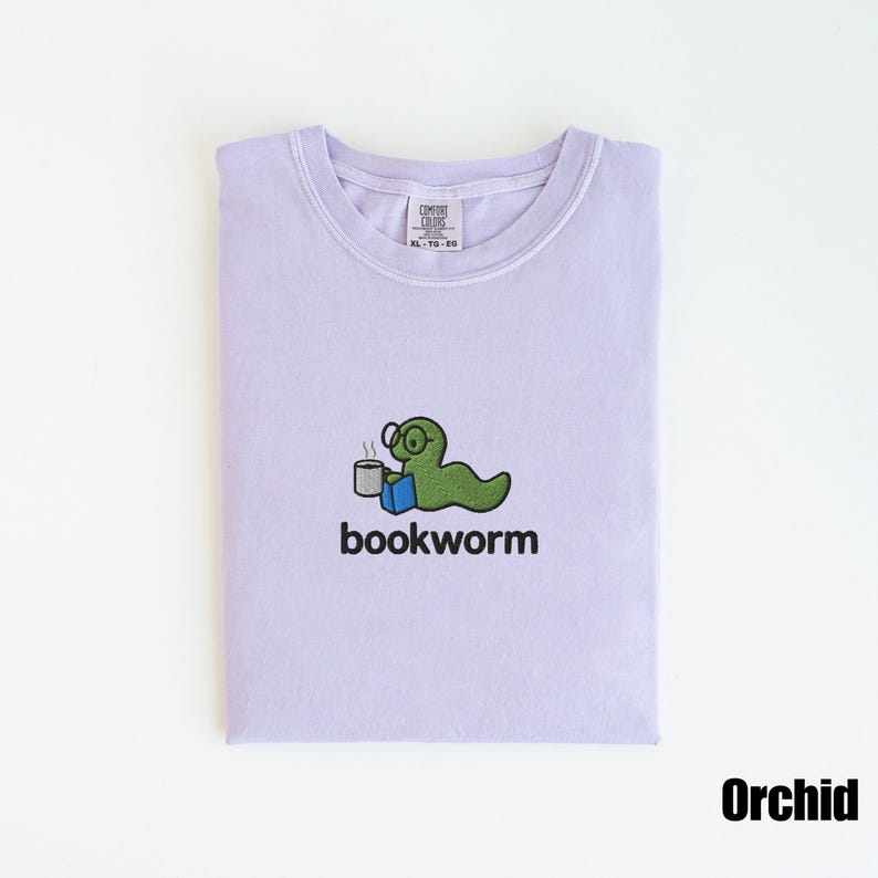 May include: A light purple t-shirt with a crew neck. The shirt features a green cartoon bookworm wearing glasses, holding a mug, and sitting on a blue book. The word "bookworm" is printed below the graphic.