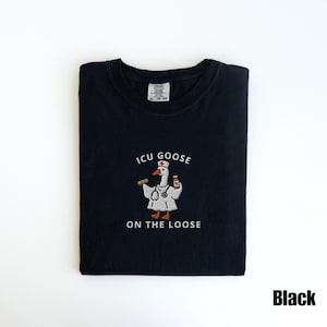 May include: Black t-shirt with the text "ICU GOOSE ON THE LOOSE" and a cartoon goose wearing a nurse's uniform. The goose is holding a stethoscope and a bottle of medicine. The shirt is folded neatly.