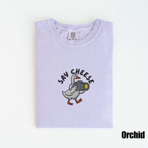 May include: Orchid-colored t-shirt with a crew neck. The shirt features an embroidered design of a goose holding a camera, with the words "SAY CHEESE" above it. The shirt is folded neatly.