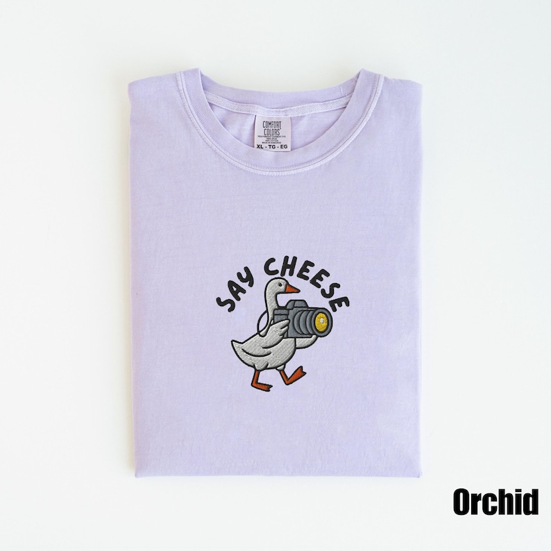 May include: Orchid-colored t-shirt with a crew neck. The shirt features an embroidered design of a goose holding a camera, with the words "SAY CHEESE" above it. The shirt is folded neatly.