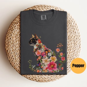 May include: A dark grey t-shirt with a floral design featuring a cat silhouette. The flowers are in shades of pink, red, white, and yellow. The text "COMFORT COLORS" is visible on the tag.