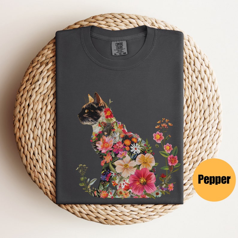 May include: A dark grey t-shirt with a floral design featuring a cat silhouette. The flowers are in shades of pink, red, white, and yellow. The text "COMFORT COLORS" is visible on the tag.