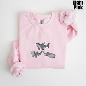 May include: Light pink sweatshirt with embroidered gray sharks. The sweatshirt features three different shark designs, including a hammerhead and a whale shark. The text "Light Pink" is in the upper right corner.