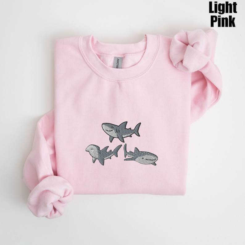 May include: Light pink sweatshirt with embroidered gray sharks. The sweatshirt features three different shark designs, including a hammerhead and a whale shark. The text "Light Pink" is in the upper right corner.
