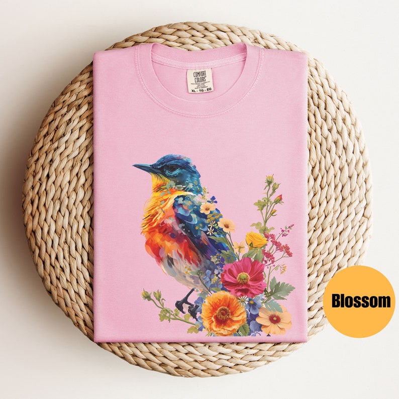 May include: Pink t-shirt featuring a colourful bird and floral design. The bird is blue, orange and yellow. The flowers are pink, yellow and blue. The shirt has the text "COMFORT COLORS" on the tag.