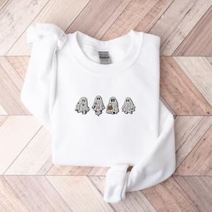 May include: A white long-sleeve crewneck sweatshirt featuring an embroidered design of four small ghosts across the chest. Each ghost figure displays a different pattern, including floral, polka dot, and pumpkin motifs in shades of orange and grey.