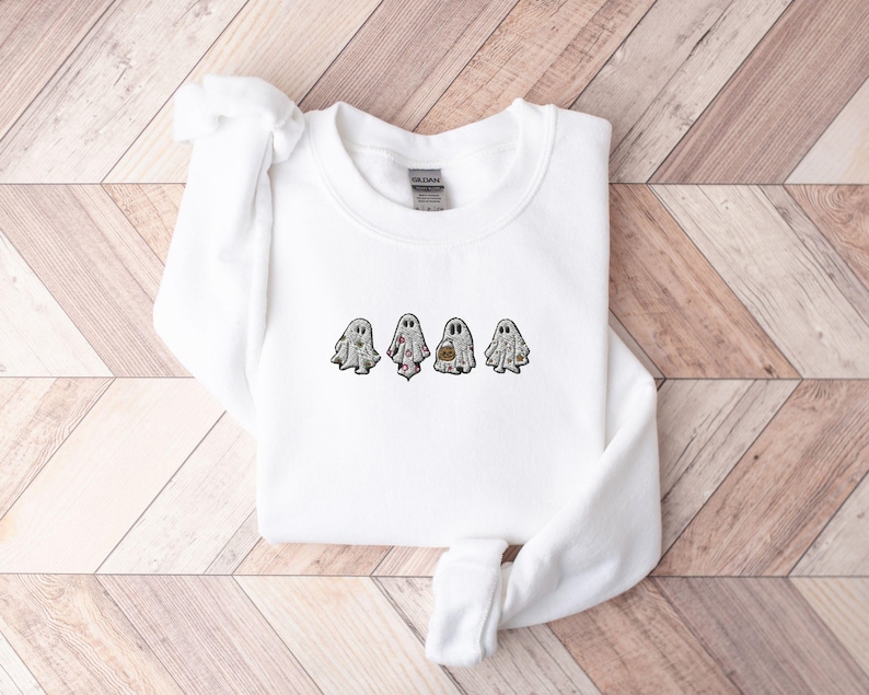 May include: A white long-sleeve crewneck sweatshirt featuring an embroidered design of four small ghosts across the chest. Each ghost figure displays a different pattern, including floral, polka dot, and pumpkin motifs in shades of orange and grey.