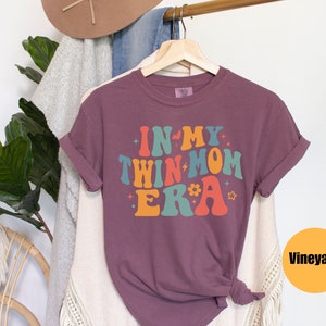 Może przedstawiać: Fioletowy T-shirt z napisem "In My Twin Mom Era" w retro czcionce tęczowej. Koszulka wisi na drewnianym wieszaku.
