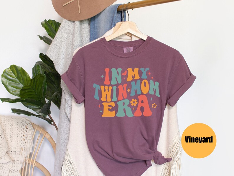 Może przedstawiać: Fioletowy T-shirt z napisem "In My Twin Mom Era" w retro czcionce tęczowej. Koszulka wisi na drewnianym wieszaku.