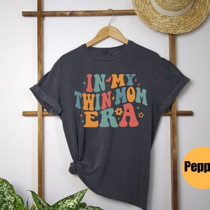 Może przedstawiać: Szary T-shirt z napisem "In My Twin Mom Era" w kolorowych, retro literach. Koszulka wisi na drewnianym stojaku.