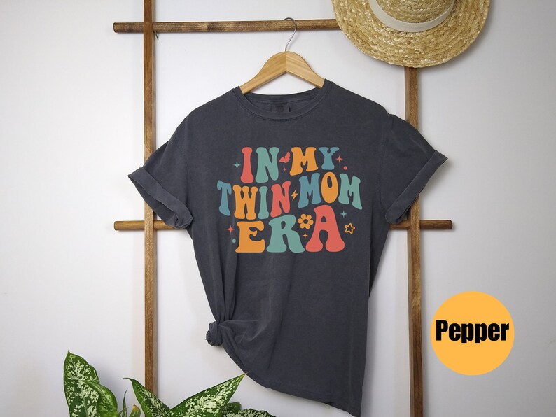 Może przedstawiać: Szary T-shirt z napisem "In My Twin Mom Era" w kolorowych, retro literach. Koszulka wisi na drewnianym stojaku.