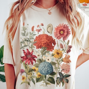 Pu&ograve; includere: Una maglietta bianca con una stampa floreale che presenta una variet&agrave; di fiori in tonalit&agrave; di rosso, rosa, giallo e blu. I fiori sono disposti in un design a bouquet e sono circondati da foglie verdi.