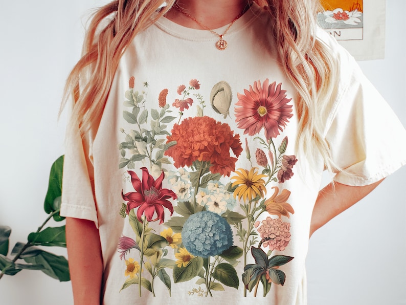 Pu&ograve; includere: Una maglietta bianca con una stampa floreale che presenta una variet&agrave; di fiori in tonalit&agrave; di rosso, rosa, giallo e blu. I fiori sono disposti in un design a bouquet e sono circondati da foglie verdi.