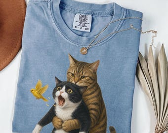 May include: A blue jean colored t-shirt featuring a graphic of a brown tabby cat hugging a black and white tuxedo cat, with a small yellow bird flying to the left. The shirt has a Comfort Colors brand label at the neck and a crew neckline.