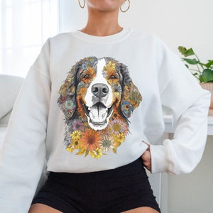 K&ouml;nnte beinhalten: Wei&szlig;er Pullover mit einem bunten Blumenmuster eines Berner Sennenhunds. Das Gesicht des Hundes ist von Blumen in Gelb-, Orange-, Rosa- und Blaut&ouml;nen umgeben.