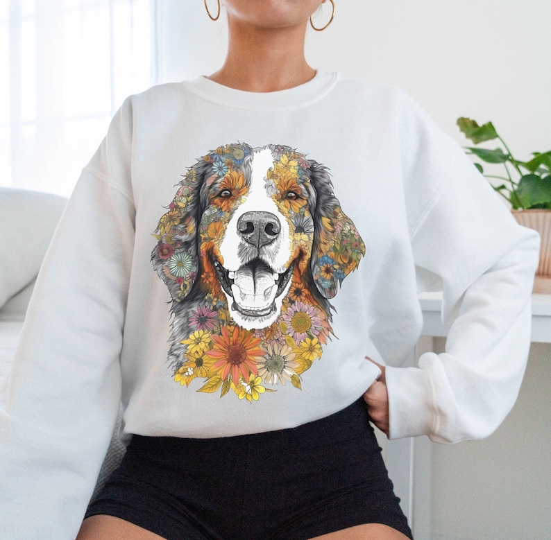K&ouml;nnte beinhalten: Wei&szlig;er Pullover mit einem bunten Blumenmuster eines Berner Sennenhunds. Das Gesicht des Hundes ist von Blumen in Gelb-, Orange-, Rosa- und Blaut&ouml;nen umgeben.