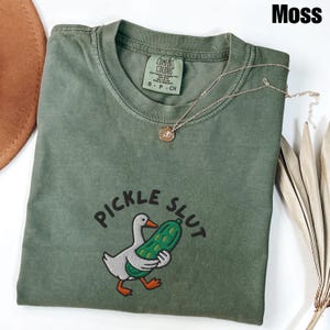 May include: A moss green t-shirt with the text "PICKLE SLUT" and an embroidered graphic of a duck holding a pickle. A gold necklace with a pendant is on the shirt. The shirt is a Comfort Colors brand, size S-P-CH.