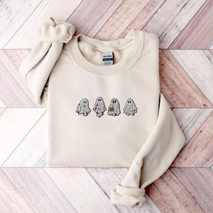 May include: A cream-coloured crewneck sweatshirt featuring an embroidered design of four small ghosts across the chest. Each ghost has a unique pattern, including floral, polka dot, and solid details, with one ghost holding a small orange pumpkin bucket.