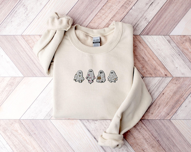 May include: A cream-coloured crewneck sweatshirt featuring an embroidered design of four small ghosts across the chest. Each ghost has a unique pattern, including floral, polka dot, and solid details, with one ghost holding a small orange pumpkin bucket.