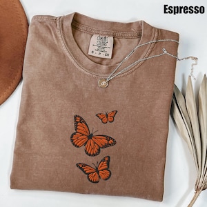 May include: Espresso-colored t-shirt with embroidered orange butterflies. The shirt has a crew neck and short sleeves. A silver necklace with a small pendant is draped across the shirt. The shirt is labeled "Comfort Colors."