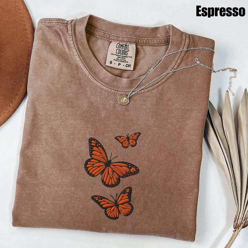 May include: Espresso-colored t-shirt with embroidered orange butterflies. The shirt has a crew neck and short sleeves. A silver necklace with a small pendant is draped across the shirt. The shirt is labeled "Comfort Colors."