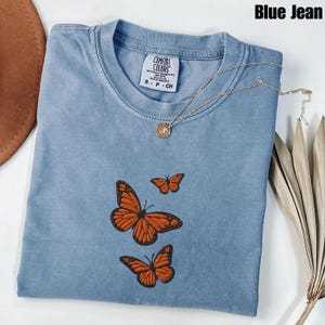 May include: A blue jean colored t-shirt with three embroidered orange butterflies. The shirt has a crew neck and short sleeves. A gold necklace with a small pendant is draped across the shirt.