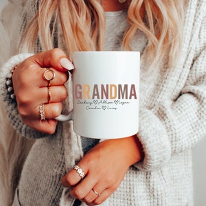 以下が含まれることがあります： 白いセラミック製のマグカップに、カラフルな文字で「Grandma」と、ハートで区切られた「Zachary、Allison、Camden、Lucas」という名前が書かれています。