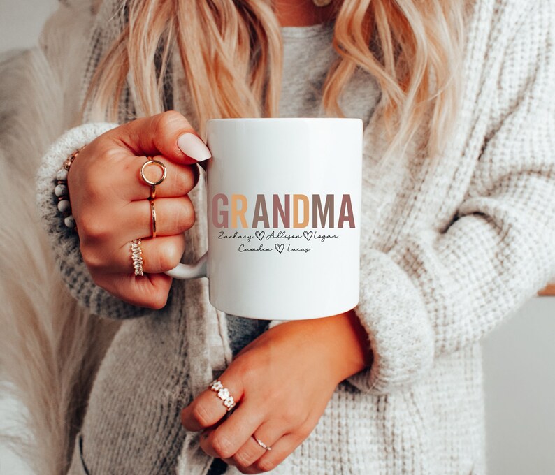 以下が含まれることがあります： 白いセラミック製のマグカップに、カラフルな文字で「Grandma」と、ハートで区切られた「Zachary、Allison、Camden、Lucas」という名前が書かれています。
