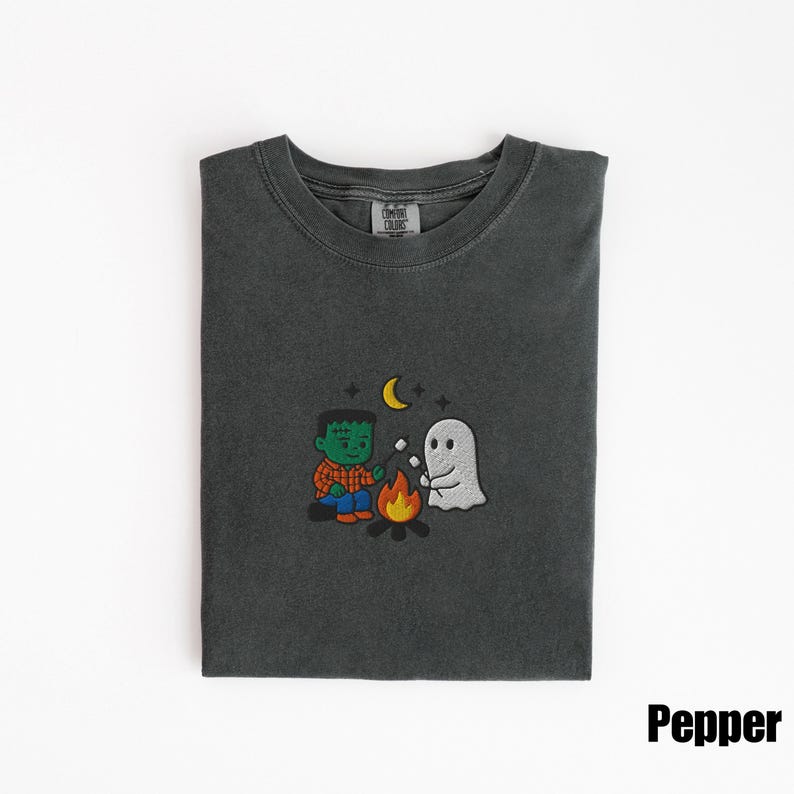May include: Dark gray t-shirt with a crew neck. The shirt features an embroidered design of a Frankenstein-like character and a ghost roasting marshmallows over a campfire, with a crescent moon in the sky. The word "Pepper" is printed on the bottom right.