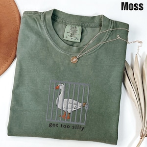 May include: Moss green t-shirt with an embroidered goose behind bars. The goose has orange feet and beak. The text "got too silly" is below the goose. A gold necklace with a pendant is on the shirt.