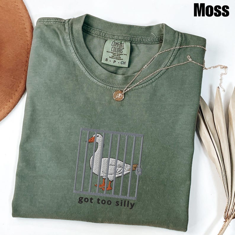 May include: Moss green t-shirt with an embroidered goose behind bars. The goose has orange feet and beak. The text "got too silly" is below the goose. A gold necklace with a pendant is on the shirt.