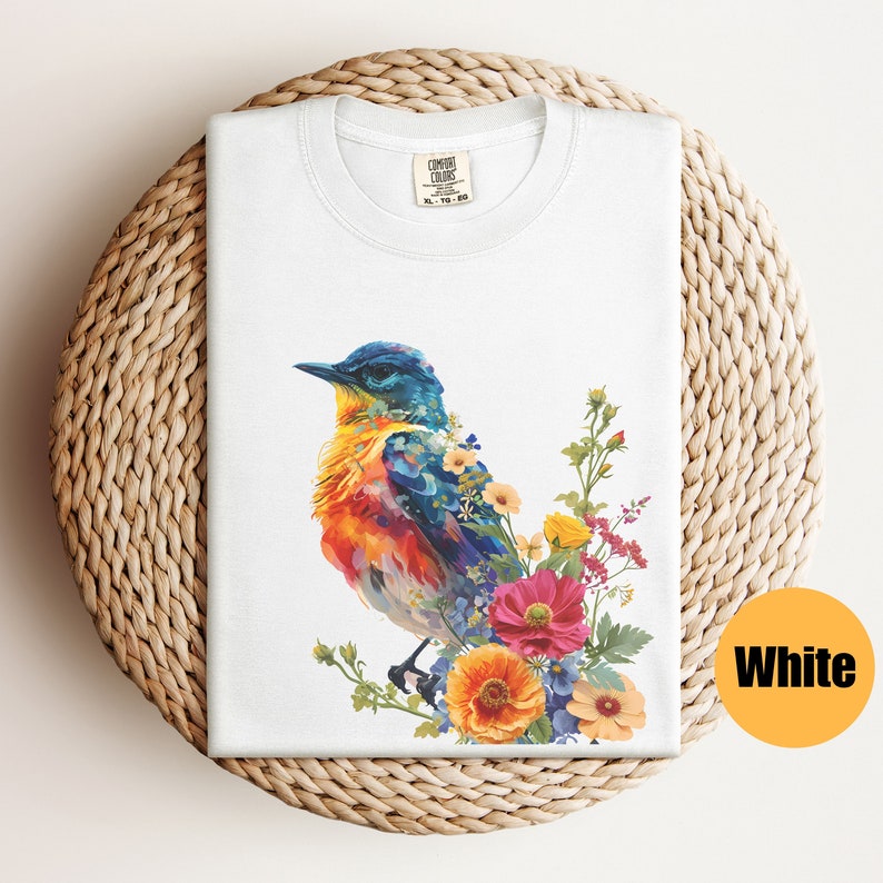 May include: White t-shirt featuring a colourful bird and floral design. The bird is blue, yellow, orange, and red. The flowers are pink, yellow, orange, and blue.