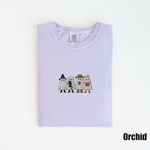 May include: A light purple t-shirt with an embroidered design of four cartoon ghosts, each wearing a different hat and holding various items. The shirt is labeled "Orchid" and has a Comfort Colors tag.