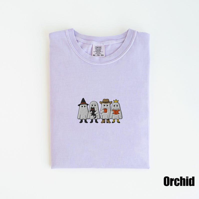 May include: A light purple t-shirt with an embroidered design of four cartoon ghosts, each wearing a different hat and holding various items. The shirt is labeled "Orchid" and has a Comfort Colors tag.