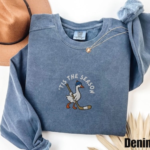 May include: Denim blue sweatshirt with a crew neck. The front features an embroidered design of a goose wearing a blue hat and holding a hockey stick, with the text "'Tis the Season" around it. A gold necklace is draped on the sweatshirt.