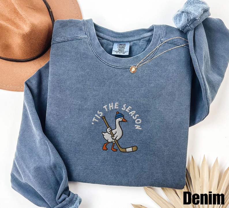 May include: Denim blue sweatshirt with a crew neck. The front features an embroidered design of a goose wearing a blue hat and holding a hockey stick, with the text "'Tis the Season" around it. A gold necklace is draped on the sweatshirt.