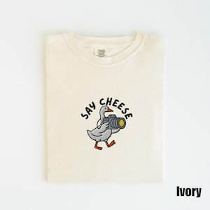 May include: Ivory-colored t-shirt featuring an embroidered design of a goose holding a camera, with the words "SAY CHEESE" above the goose. The shirt is folded neatly. The word "Ivory" is printed in black at the bottom right.