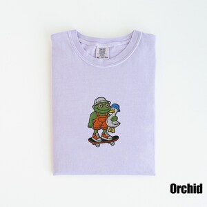 May include: Orchid-colored t-shirt with an embroidered design of a frog wearing overalls, a hat, and sunglasses, skateboarding with a duck. The shirt has a crew neck and a tag that reads "Comfort Colors". The word "Orchid" is printed at the bottom.